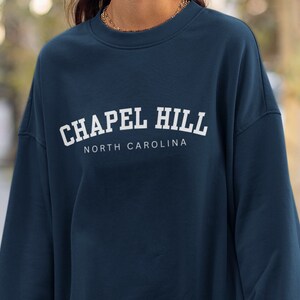Puede incluir: Una sudadera azul oscuro con texto blanco que dice "Chapel Hill North Carolina".