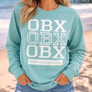 May include: A teal long-sleeve sweatshirt with the text "OBX" stacked three times in white, with "NORTH CAROLINA" below. The sweatshirt is a solid teal color and has a crew neck.