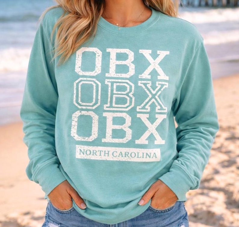 Puede incluir: Una sudadera de manga larga de color verde azulado con el texto "OBX" apilado tres veces en blanco, con "NORTH CAROLINA" debajo. La sudadera es de color verde azulado liso y tiene cuello redondo.