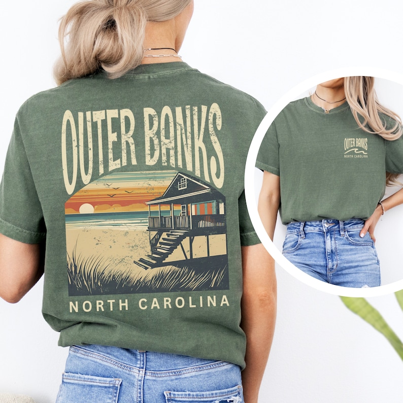 May include: Olive green t-shirt featuring a beach house graphic with the text "Outer Banks North Carolina." The design includes a sunset, ocean, and beach house. The shirt is a short-sleeved crew neck.