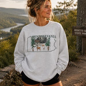 Peut inclure: Un sweat-shirt gris clair &agrave; col rond arborant le contour noir de l'&Eacute;tat de Pennsylvanie avec le mot PENNSYLVANIA imprim&eacute; au-dessus. &Agrave; l'int&eacute;rieur du contour se trouve une illustration graphique d'un chalet, de sapins et de plusieurs cerfs.