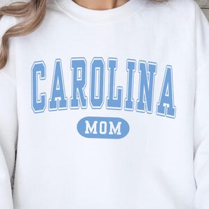 Puede incluir: Sudadera blanca con la palabra "CAROLINA" en letras de bloque azul claro, con un contorno azul claro. Debajo de la palabra "CAROLINA" est&aacute; la palabra "MOM" en un &oacute;valo azul claro.
