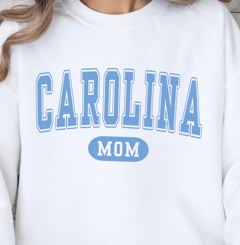Puede incluir: Sudadera blanca con la palabra "CAROLINA" en letras de bloque azul claro, con un contorno azul claro. Debajo de la palabra "CAROLINA" est&aacute; la palabra "MOM" en un &oacute;valo azul claro.