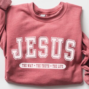 May include: A heathered red crewneck sweatshirt featuring the word JESUS in large white block letters with a distressed texture. Below the main text, a white horizontal bar contains the words THE WAY &bull; THE TRUTH &bull; THE LIFE in red.