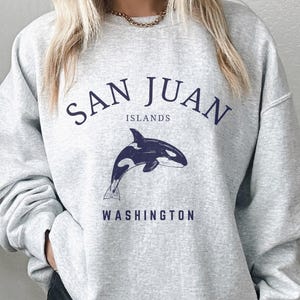 May include: A light grey crewneck sweatshirt featuring dark blue text that reads SAN JUAN ISLANDS WASHINGTON and a graphic illustration of an orca whale centred on the chest. The long-sleeved garment has a ribbed neckline and cuffs.