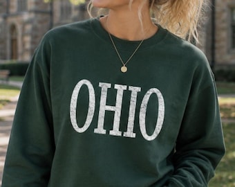 May include: A dark forest green crewneck sweatshirt featuring the word OHIO printed in large, white, distressed-style block lettering across the chest. The garment has long sleeves and a ribbed neckline, providing a simple and classic appearance.