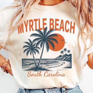 May include: A cream-colored t-shirt featuring a graphic print with the text MYRTLE BEACH in orange letters at the top and South Carolina in orange script at the bottom. The design includes dark blue palm trees, a large orange sun, and a beach scene.
