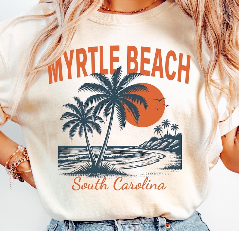 May include: A cream-colored t-shirt featuring a graphic print with the text MYRTLE BEACH in orange letters at the top and South Carolina in orange script at the bottom. The design includes dark blue palm trees, a large orange sun, and a beach scene.