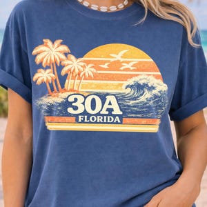 May include: Navy blue t-shirt with a sunset graphic, palm trees, waves, and the text "30A FLORIDA". The design uses orange, yellow, and white. The shirt has rolled-up sleeves.