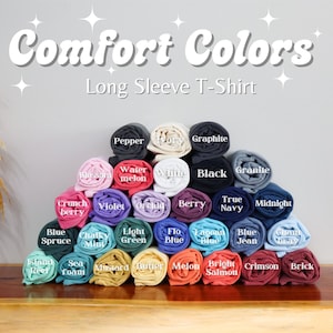 Puede incluir: Una exhibici&oacute;n de camisetas de manga larga enrolladas en varios colores, incluyendo negro, azul y rosa. El texto "Comfort Colors" y "Long Sleeve T-Shirt" est&aacute; en la parte superior. Las camisetas est&aacute;n dispuestas en forma de pir&aacute;mide, mostrando una gama de tonos.