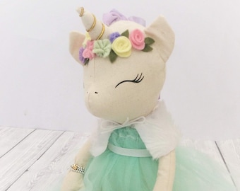 May include: A white unicorn doll wearing a green tulle dress with a white fur trim jacket and a flower crown. The doll has a gold horn and is wearing white shoes.