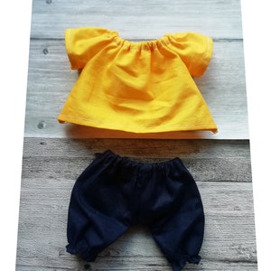 Puede incluir: Un conjunto de mu&ntilde;eca con una blusa amarilla con costuras blancas y mangas cortas, combinada con unos bombachos azul oscuro. La ropa de mu&ntilde;eca es de tela y se exhibe sobre una superficie de madera.