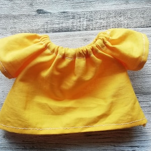 Puede incluir: Una blusa amarilla brillante, de manga corta, con cuello y mangas fruncidos. Los bordes est&aacute;n acabados con costuras blancas. La blusa est&aacute; hecha de una tela suave.