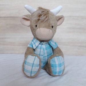 Puede incluir: Un peluche de vaca de las Tierras Altas con un cuerpo marrón claro, cuernos blancos y un atuendo a cuadros azul y blanco. El juguete tiene un aspecto suave y es adecuado para niños.