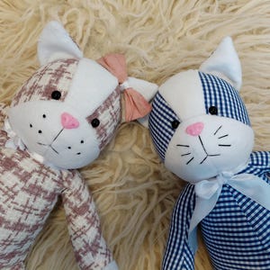 May include: Two plush cat dolls with white and patterned fabric. One cat has a pink bow and a pink nose, the other has a blue and white gingham pattern and a white bow. Both have black eyes and whiskers.