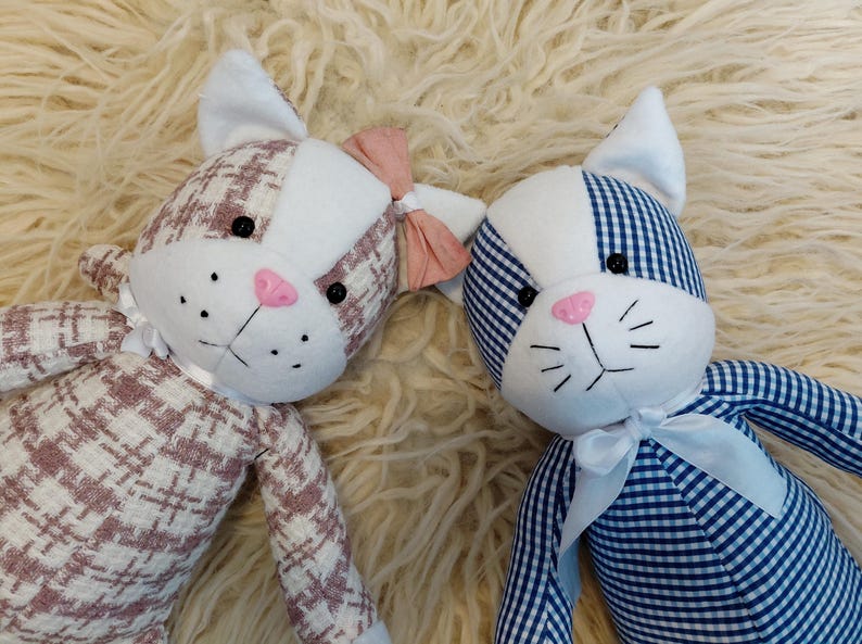 May include: Two plush cat dolls with white and patterned fabric. One cat has a pink bow and a pink nose, the other has a blue and white gingham pattern and a white bow. Both have black eyes and whiskers.
