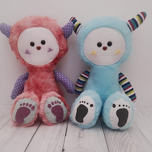 May include: Two plush toys, one pink and one blue, with smiling faces and large feet. They have horns and are made of soft, fuzzy fabric.