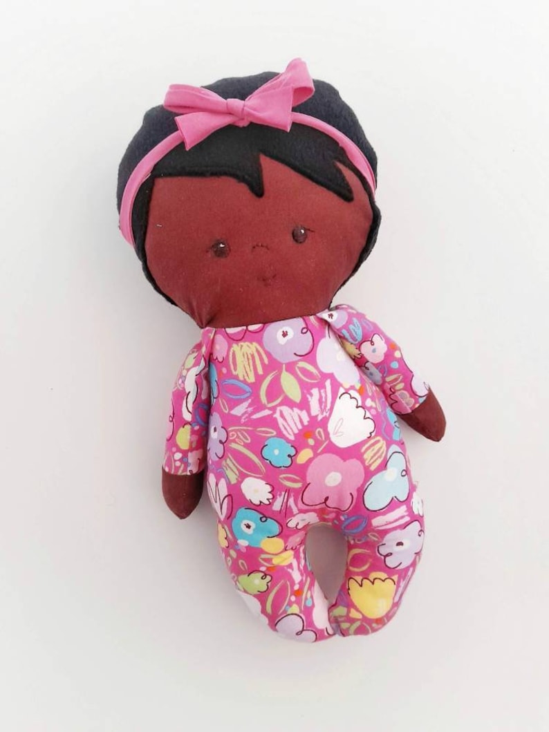 May include: A handmade doll with brown skin, black hair, and a pink bow. The doll is wearing a pink and white floral print outfit.