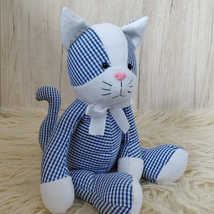 May include: A stuffed cat toy with a blue and white gingham pattern. The cat has white paws, ears, and a face with a pink nose and black eyes. A white bow is tied around its neck. The cat is sitting on a fluffy, cream-colored surface.