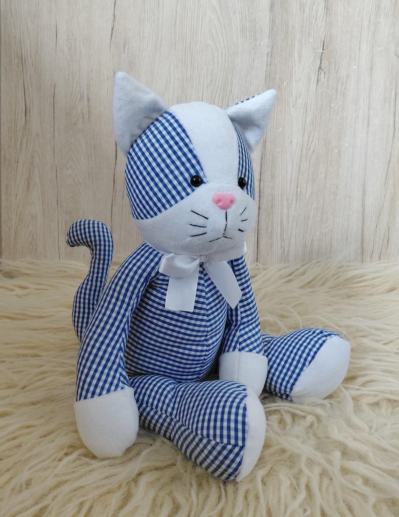 May include: A stuffed cat toy with a blue and white gingham pattern. The cat has white paws, ears, and a face with a pink nose and black eyes. A white bow is tied around its neck. The cat is sitting on a fluffy, cream-colored surface.