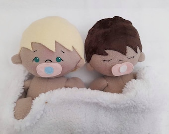May include: Two plush baby dolls, one with blonde hair and one with brown hair, are lying side-by-side in a white blanket. Both dolls have pacifiers in their mouths.