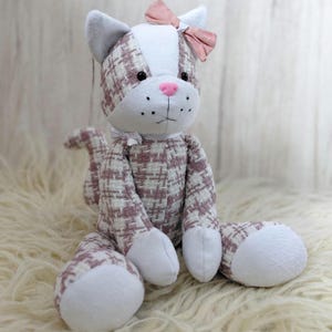 May include: A plush cat toy with a pink bow, white paws, and a pink nose. The cat's body is patterned with a white and pink plaid design. The cat is sitting on a fluffy, white surface.