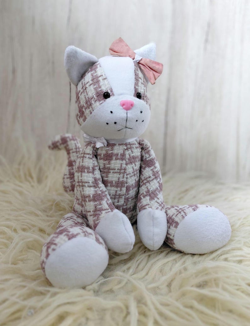 May include: A plush cat toy with a pink bow, white paws, and a pink nose. The cat's body is patterned with a white and pink plaid design. The cat is sitting on a fluffy, white surface.