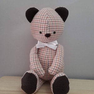 Könnte beinhalten: Ein Teddybär aus kariertem Stoff mit braunen Ohren und Füßen. Der Bär trägt eine weiße Schleife.