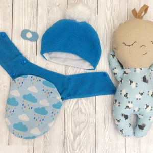 May include: A handmade baby doll with a blue and white sheep print onesie, a blue fleece hat with a white pom-pom, and a blue fleece bib with a cloud and raindrop print.