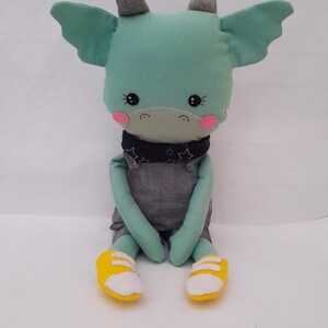 May include: A teal blue plush dragon toy wearing a black and white bandana with stars and gray overalls. The dragon has pink cheeks and black eyes. The dragon is wearing yellow shoes with white laces.