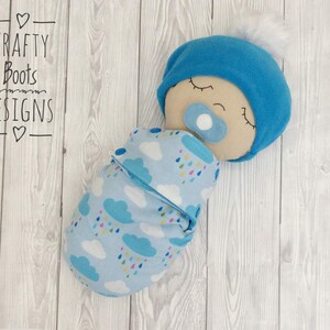 May include: A blue and white fabric baby doll with a blue hat and a pacifier. The doll is wrapped in a blue and white blanket with a cloud and raindrop pattern. The doll is lying on a white wooden surface.