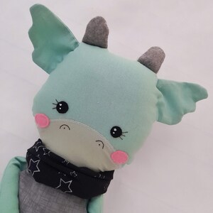 May include: A light blue plush toy dragon with gray horns and wings. The dragon has black eyes, pink cheeks, and a black and white star patterned bandana.