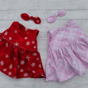 May include: Two sleeveless dresses with matching hair bows. One dress is red with a white snowflake pattern, and the other is a light pink and white plaid pattern. Both garments feature a gathered waistline and a simple round neckline design.