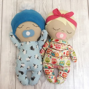 May include: Two handmade cloth baby dolls with closed eyes and pacifiers. The doll on the left is wearing a blue hat and blue and white pajamas with a sheep pattern. The doll on the right is wearing a pink headband and a white and brown pajama with an owl pattern.