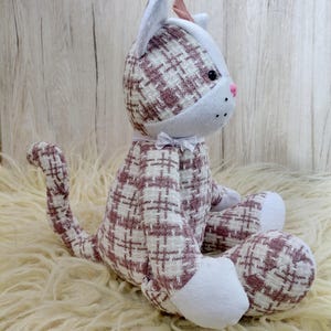 May include: A plush cat doll with a white and mauve plaid pattern. The cat has white paws, ears, and a face, with a pink nose and a small bow. The cat is sitting on a cream-colored, fluffy surface.