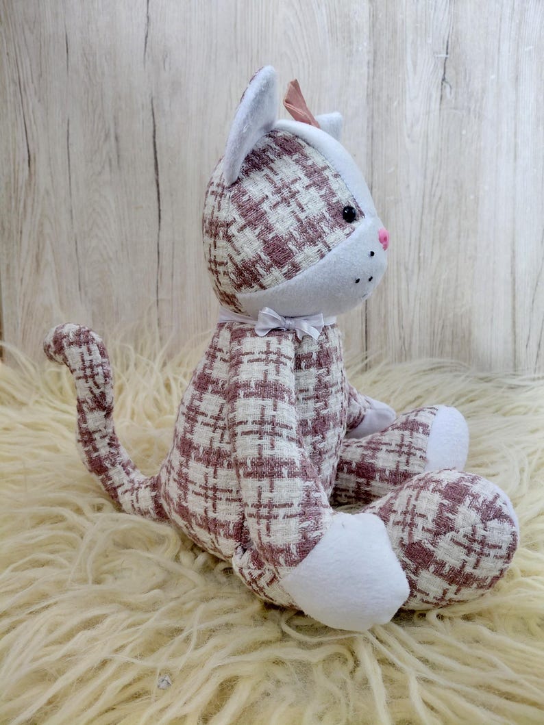 May include: A plush cat doll with a white and mauve plaid pattern. The cat has white paws, ears, and a face, with a pink nose and a small bow. The cat is sitting on a cream-colored, fluffy surface.