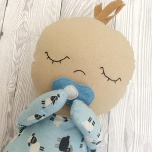 May include: A plush toy baby doll with a pacifier, closed eyes, and a brown hair tuft. The doll is wearing blue pajamas with a white sheep pattern.