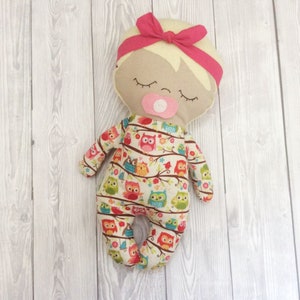 May include: A handmade fabric doll with a pink headband, closed eyes, and a pacifier. The doll is wearing a white onesie with a colorful owl print.