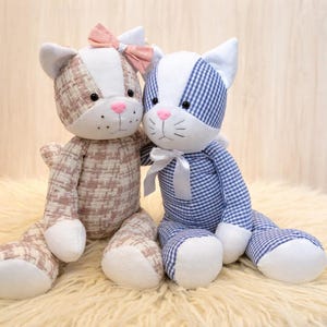 May include: Two plush cat dolls. One cat is patterned in pink and white plaid with a pink bow. The other cat is blue and white gingham with a white bow. Both have pink noses and white paws.
