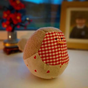 May include: A handmade fabric bird with a burlap head and a red and white checkered breast. The bird's belly is a light beige fabric with small red hearts. It has a small orange beak and black eyes.
