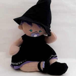 May include: A plush witch doll wearing a black hat and dress with a purple bow. The doll has a dummy in its mouth and is sitting on a white surface.