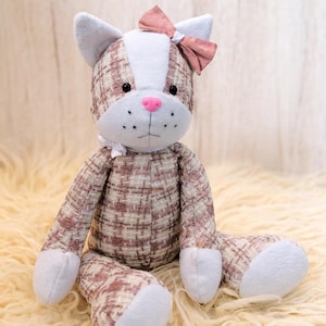 May include: A plush cat doll with a pink bow, pink nose, and black eyes. The cat's body is made of a brown and white plaid fabric, with light blue accents on the ears, paws, and face. The doll is sitting on a fluffy, cream-colored surface.