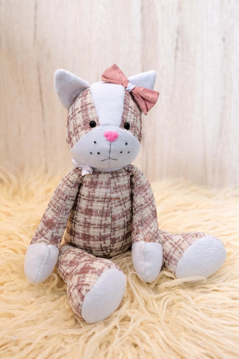 May include: A plush cat doll with a pink bow, pink nose, and black eyes. The cat's body is made of a brown and white plaid fabric, with light blue accents on the ears, paws, and face. The doll is sitting on a fluffy, cream-colored surface.