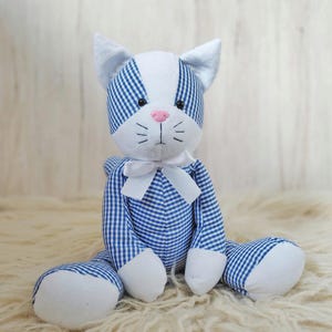 May include: A stuffed cat toy with a blue and white gingham pattern. The cat has white paws, ears, and face, a pink nose, and a white bow tie. The toy is sitting on a fluffy, cream-colored surface.
