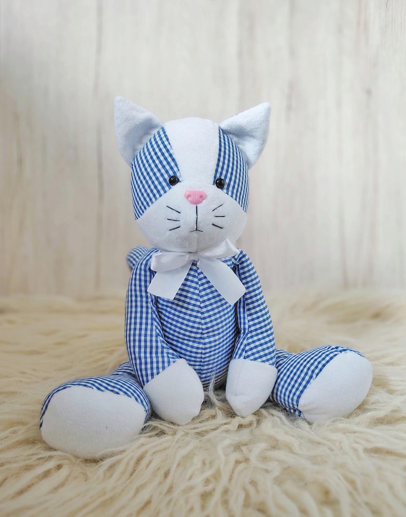 May include: A stuffed cat toy with a blue and white gingham pattern. The cat has white paws, ears, and face, a pink nose, and a white bow tie. The toy is sitting on a fluffy, cream-colored surface.