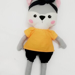 Puede incluir: Una mu&ntilde;eca de lobo de peluche hecha a mano con pelaje gris, orejas internas rosas y una diadema negra. La mu&ntilde;eca lleva una camisa naranja, pantalones cortos negros y zapatos blancos. La cara es blanca con ojos negros y nariz negra.