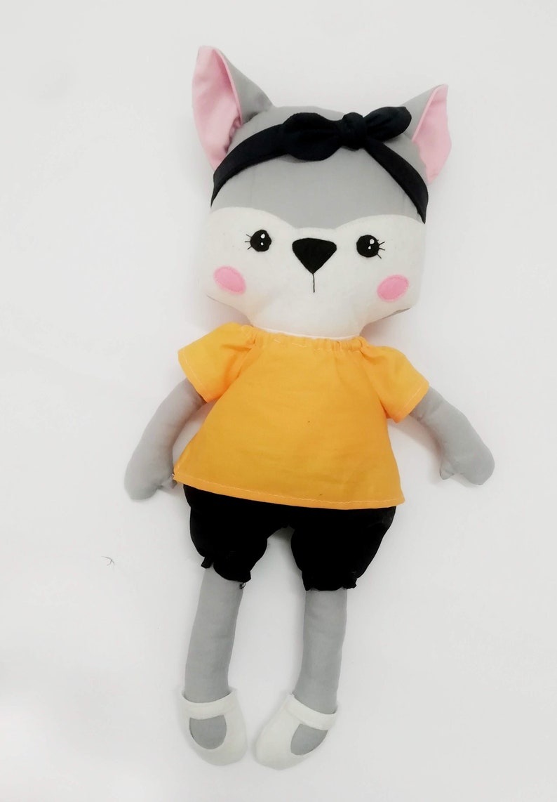 Puede incluir: Una mu&ntilde;eca de lobo de peluche hecha a mano con pelaje gris, orejas internas rosas y una diadema negra. La mu&ntilde;eca lleva una camisa naranja, pantalones cortos negros y zapatos blancos. La cara es blanca con ojos negros y nariz negra.