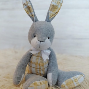May include: A grey plush rabbit with long ears and a white bow. The rabbit's ears, belly, and feet have a yellow and white plaid pattern. The soft toy is sitting on a cream-coloured, fluffy surface.