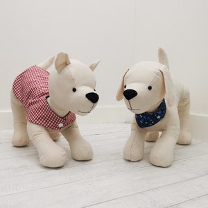 Puede incluir: Dos figuras de perro de peluche. Uno lleva una camisa a cuadros rojos y blancos, el otro una bandana azul con estrellas blancas. Ambos tienen narices y ojos negros y están a cuatro patas.