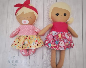 May include: Two handmade fabric dolls with blonde hair and painted faces. One doll is wearing a pink and yellow dress with a pacifier. The other doll is wearing a pink dress with a floral pattern. The dolls are sitting on a white wooden surface. Crafty Boots Designs.